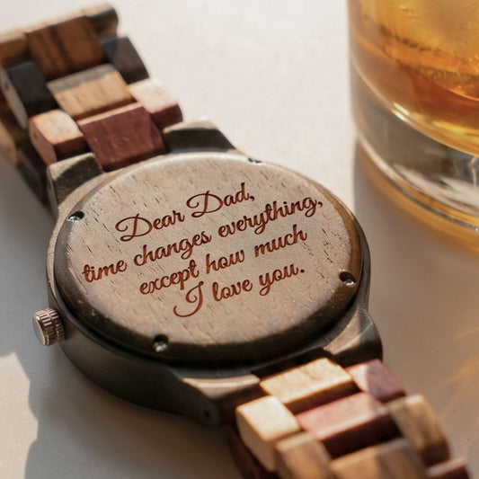 A wooden link watch shown from the back with an engraved message for Dad, placed next to a glass of whiskey on a light surface.