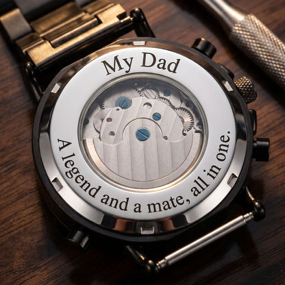 A close-up of a stainless steel wood watch back with engraved text reading 'My Dad' and 'A legend and a mate, all in one.' The transparent center reveals the mechanical movement, and the watch is placed on a dark wooden surface.