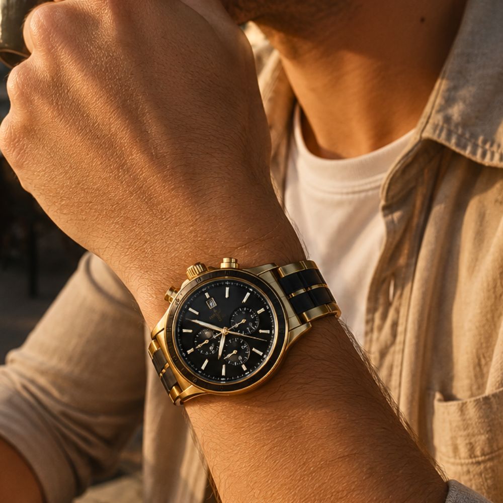 A close-up lifestyle shot of a man wearing a gold-tone chronograph watch with a black dial and a black-and-gold striped metal bracelet, set against a warm, golden-hour background.